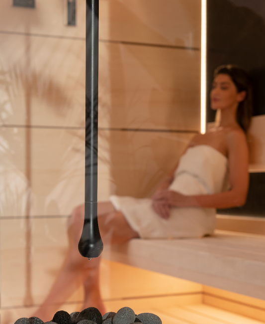 Person sitting in a sauna with a black handheld device, surrounded by stones.
