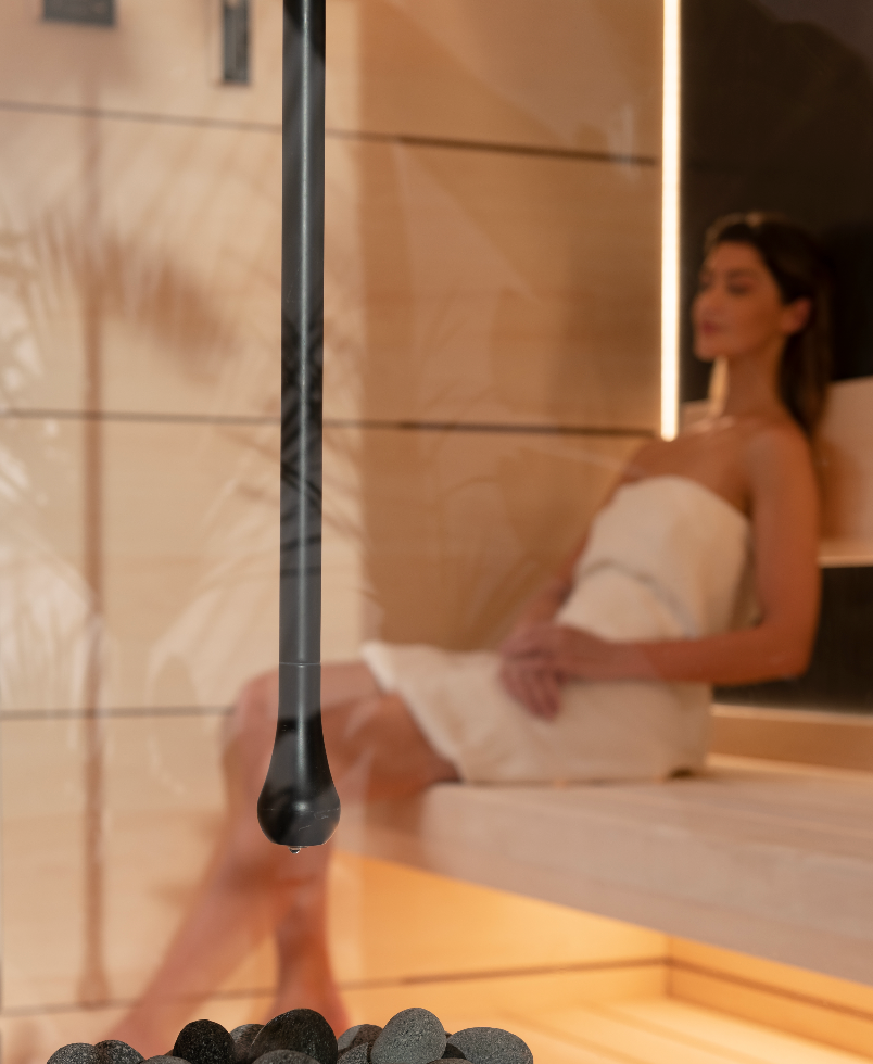 Person sitting in a sauna with a black handheld device, surrounded by stones.