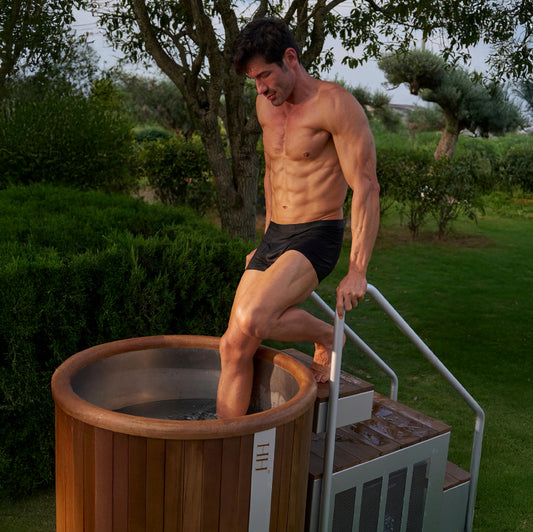 Man stepping into a luxury wooden cold plunge tub with stainless steel interior and side stairs, surrounded by greenery, showcasing the Haus of Nord by Haus of Heat.