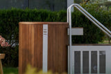 A luxury outdoor cold plunge tub made of walnut wood and stainless steel, featuring side stairs and the Haus of Heat logo, set in a serene garden environment.