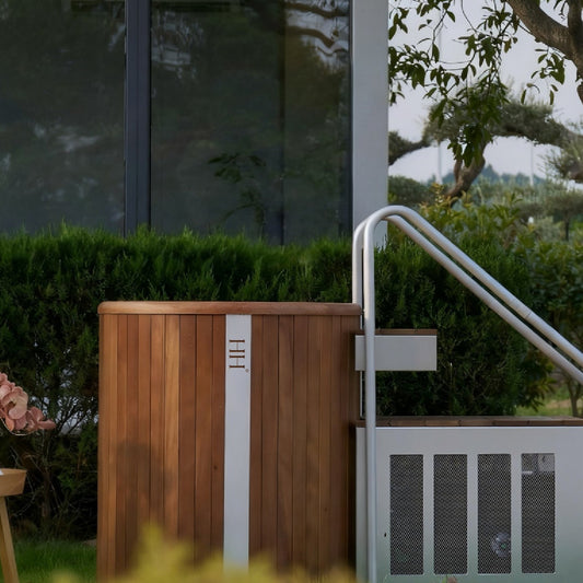 A luxury outdoor cold plunge tub made of walnut wood and stainless steel, featuring side stairs and the Haus of Heat logo, set in a serene garden environment.