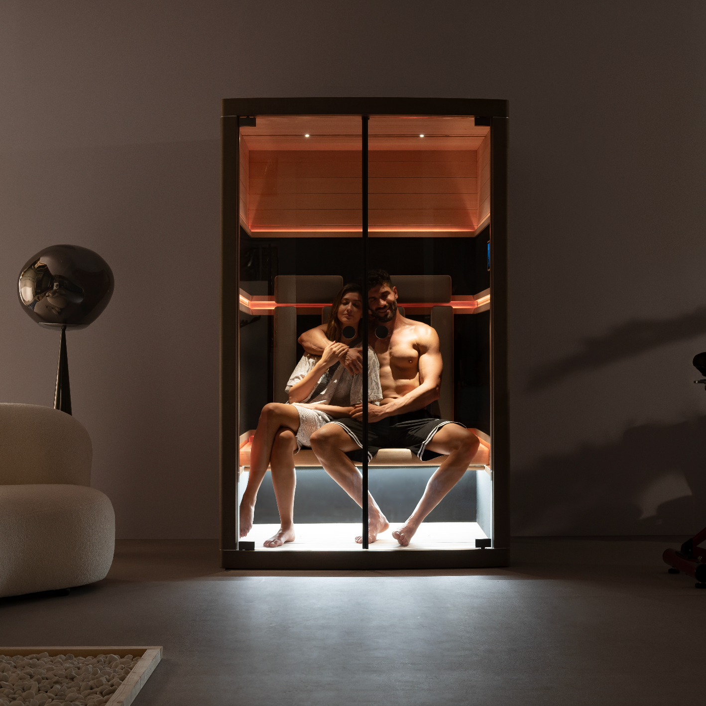 Two people sitting inside a sauna in a dark room with minimal furniture.