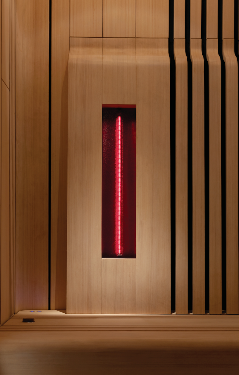 A modern sauna interior with light wood paneling, a slatted side wall, and a central red infrared light tube glowing vertically.
